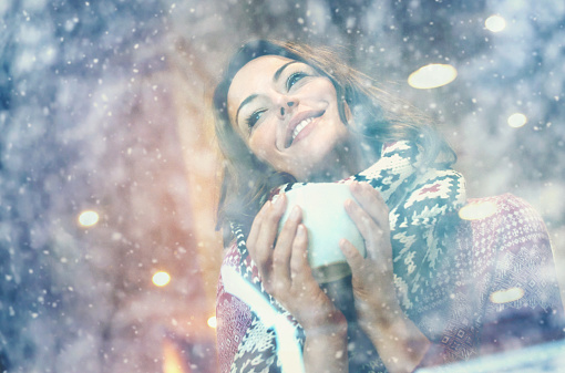 Closeup of woman having a cup of tea or coffee on snowy day. She's sitting in a cafe and looking through window. Wearing knitted cardigan and scarf and enjoying winter in warm and cosy place. Low angle shot. Visible reflection in the window.