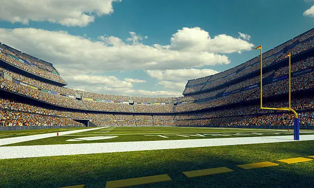 A wide angle panoramic image of a outdoor american football stadium full of spectators under blue sky. The image has depth of field with the focus on the foreground part of the pitch. The view from back line of the field.