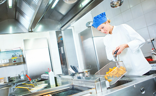 Closeup of mid 30's female chef frying some chunky potatoes in large restaurant fryer. She's actually draining the potatoes from excess fat. Large stainless steel counters, stoves and freezers around her. She's wearing chef's whites and blue chef's hat.