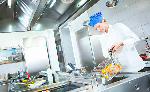 Female chef frying potatoes in restaurant kitchen. Closeup of mid 30's female chef frying some chunky potatoes in large restaurant fryer. She's actually draining the potatoes from excess fat. Large stainless steel counters, stoves and freezers around her. She's wearing chef's whites and blue chef's hat. meat locker photos stock pictures, royalty-free photos & images
