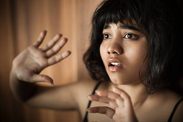 Frightened teenage girl looking away and hiding herself behind hands. Indoor close-up head and shoulder low key image shot by using single light of a lonely, cute, frightened Asian, Indian girl looking away at something and hiding herself behind her hands. She has short hair and wearing a black spaghetti strap top. Horizontal composition with copy space and selective focus. domestic violence india stock pictures, royalty-free photos & images