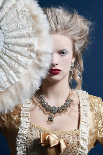 Retro baroque fashion woman wearing gold dress. Holding a fan. Studio shot against blue.