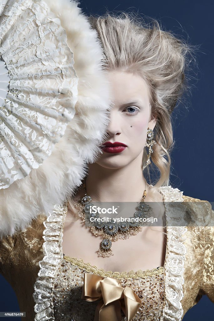 Retro baroque fashion woman wearing gold dress. Holding a fan. Retro baroque fashion woman wearing gold dress. Holding a fan. Studio shot against blue. Baroque Style Stock Photo