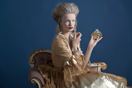 Retro baroque fashion woman wearing gold dress. Holding bottle of parfume. Sitting on vintage couch. Studio shot against blue.