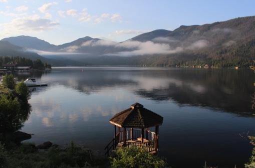 Early morning sun illuminates a hexagonal-shaped cedar Gazebo, mist clouds, shoreline, boats, and mountains at Grand Lake, Colorado