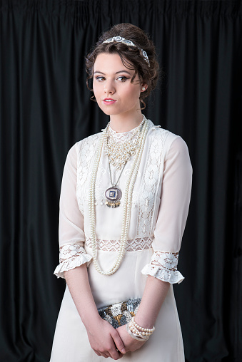 Portrait of beautiful Caucasian female model wearing Victorian style white dress and jewellery against black backdrop