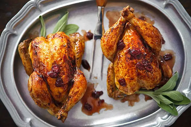 Pair of cornish game hens roasted in cranberry sauce served with fresh sage on a pewter platter.