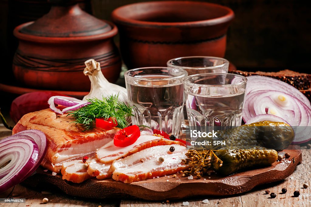 Russian tradition: cold vodka and a snack of bacon, pickles Russian tradition: cold vodka and a snack of bacon, pickles, red onion and black bread on a wooden cutting board in a rustic style, selective focus 2015 Stock Photo