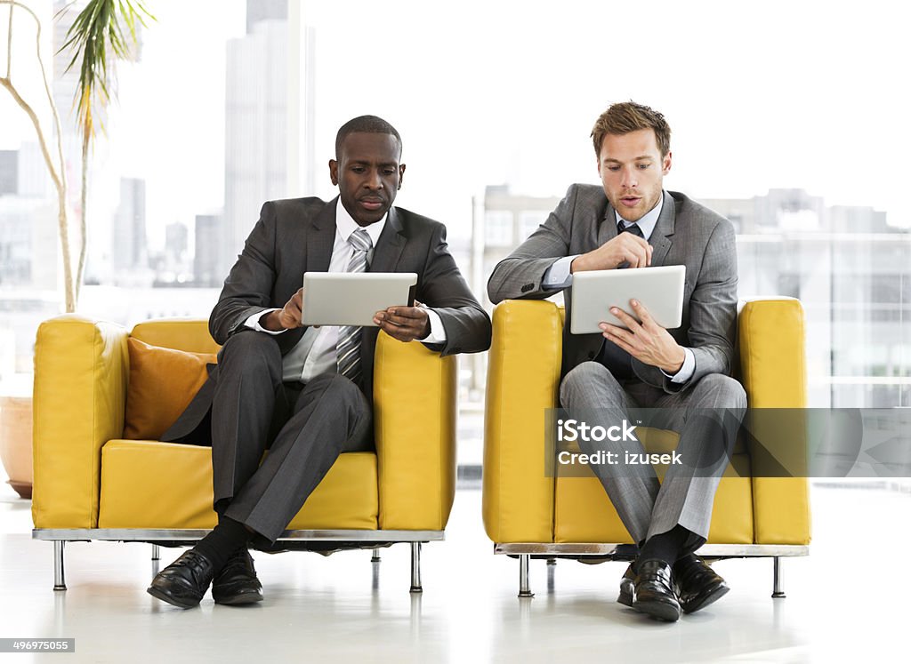 Two businessmen with digital tablets Two businessmen sitting in a chair in an office and working on digital tablets together. Office Stock Photo