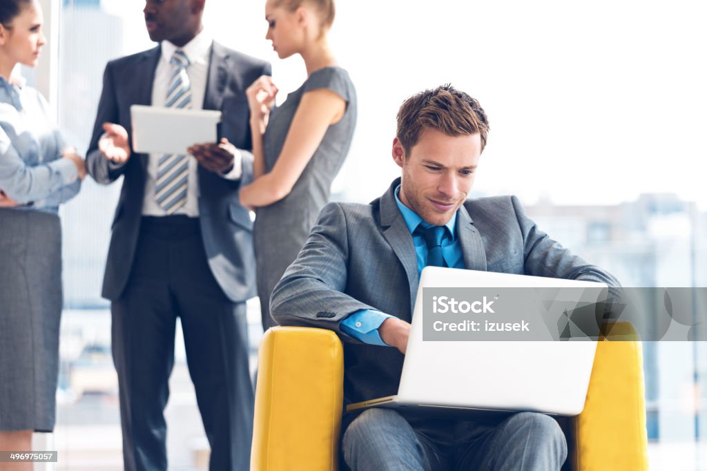 Businessman with laptop Focus on the businessman sitting in a chair and using a laptop with a group business people talking in the background. Occupation Stock Photo
