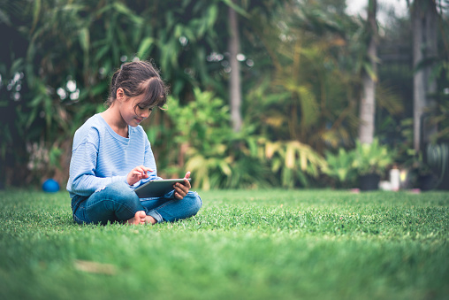 Young girl reading on digital tablet. She is sitting on the grass on the backyard, looking at tablet and smiling.