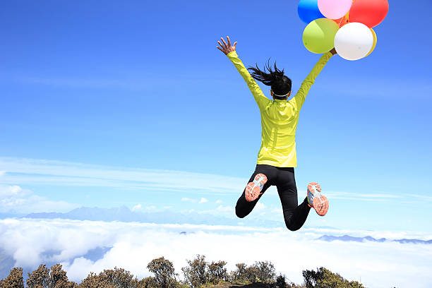 cheering young woman jumping on mountain peak with colorful balloons