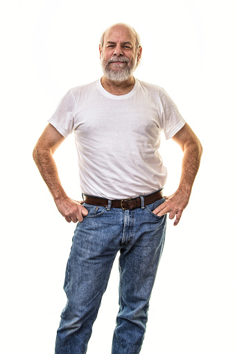 A relaxed, smiling 60-something grey beard senior adult man is standing facing the camera in a confident pose with his hands on his hips. He is wearing a white t-shirt and blue jeans. An old bald guy dressed in retro Fifties fashion style.