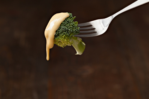 A close up horizontal photograph of a fork with a bite of broccoli topped with golden cheese sauce. Isolated on black with ample copy space.