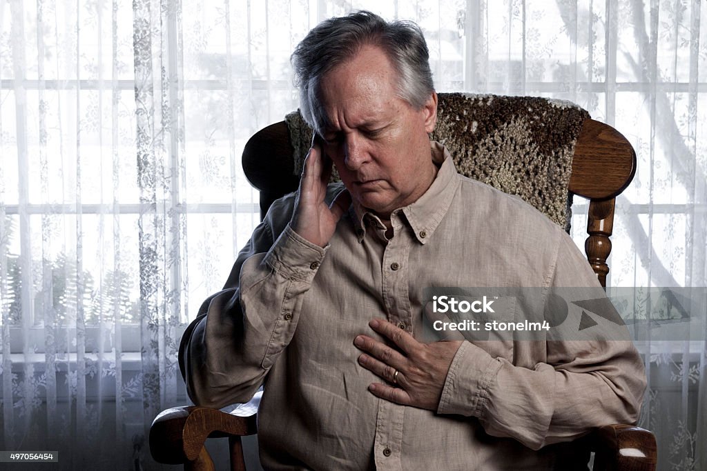 Man Suffering with Chest and Head Pain Older man sitting in a rocking chair and wincing holding his chest and head Cardiomyopathy Stock Photo