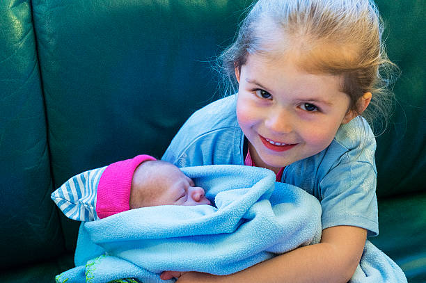 Siblings, elder sister is holding her little brother on hands. Girl is sitting on the sofa, baby boy is wrapped in a blue blanket.