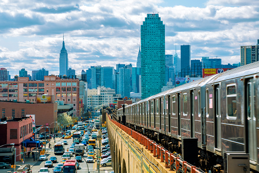 Subway train speeding on elevated track in Queens, New York. Busy street with traffic is visible down on the left side. Manhattan's finanancial and residential buildings with Empire State Building are seen in the background, horizontal orientation, USA