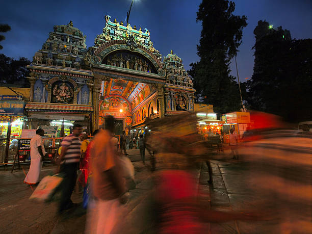 Meenakshi Temple It is evening/night time in front of exit gate with fence of Meenakshi Temple.  Blurred people are walking in front of temple while are some waiting in front the temple or looking the paintings in temple gate. On sides of gate are some shops. In background are trees, light and dark blue sky. It is in camera HDR in Madurai, Tamil Nadu, India.  menakshi stock pictures, royalty-free photos & images