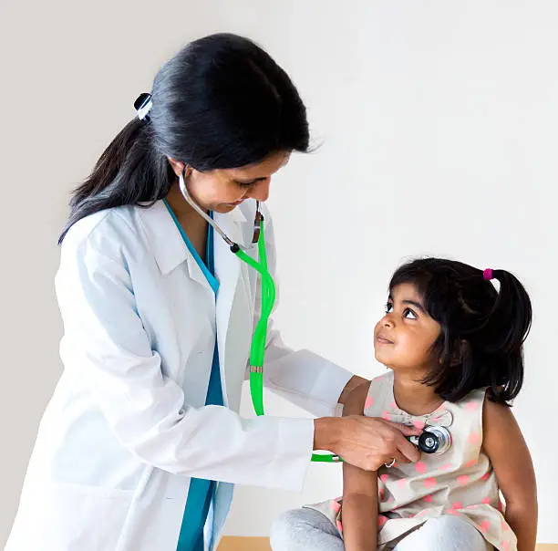 Female pediatrician checking child's lungs with stethoscope. Little girl is 5-6 years old and looking up at the doctor. Both are smiling at each other. Physician is wearing a white lab coat. Girl and doctor are of Indian ethnicity.   