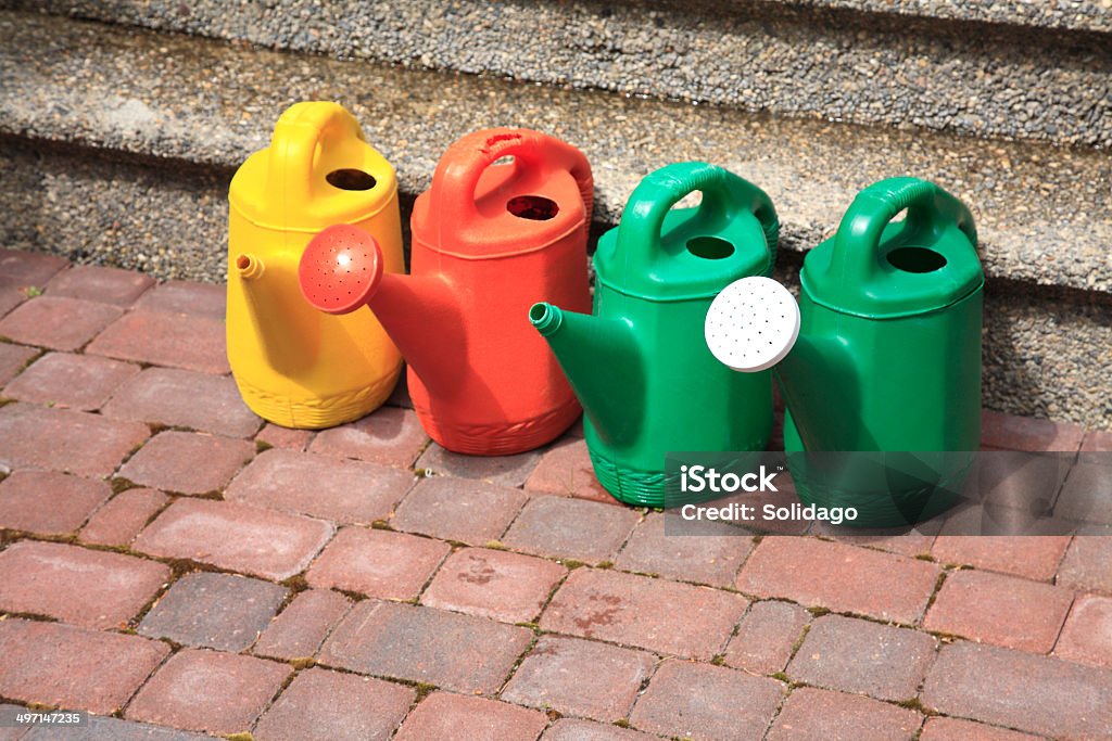 Watering Cans Ready For Service Four plastic watering cans lined up on brick patio.  Yellow,red and green. Brick Stock Photo