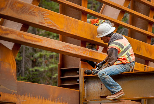 An Hispanic steel worker working high up on a girder. He is standing on the girder, wearing a safety harness, reaching up for a roof joist or truss which is being lifted into place by a crane. He has a welding helmet on his head.