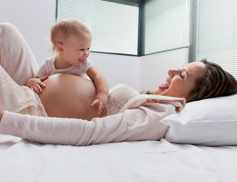 Young mother in advanced stages of pregnancy lying on bed with her infant child as the two joyfully bond with her unborn foetus as she allows the toddler to feel the baby kicking in her belly.