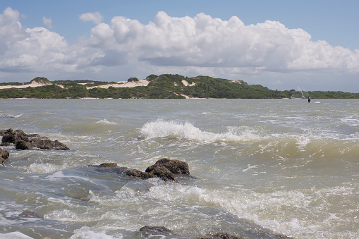 Blue sky with white puffy (Cumulus) clouds, a stretch of Brazilian coastline, seawater with waves, rocky shore and (at a distance) a fisherman casting his net into the sea to catch his dinner.