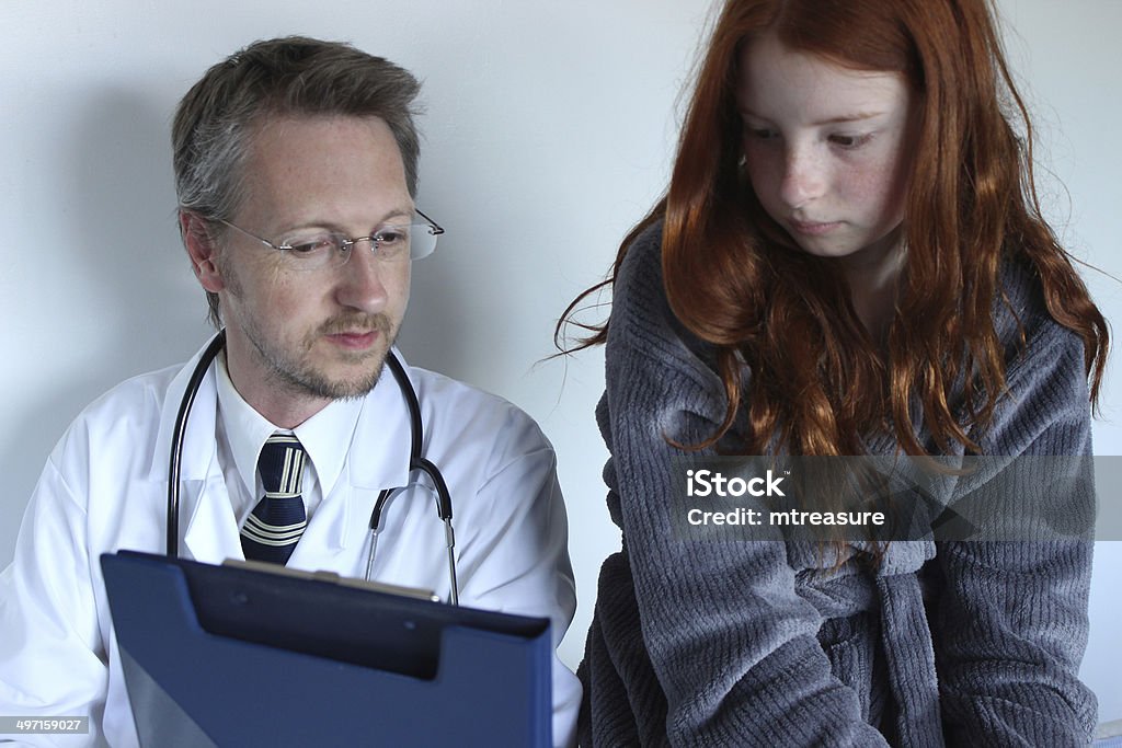 Hospital doctor showing notes to girl patient on his clipboard Photo showing a young hospital doctor showing his notes to his patient, a poorly girl.  The girl is pictured sitting on his examination table / bed, wearing her grey dressing gown and talking to the doctor as she explains her symptoms and he discussed his diagnosis. Accidents and Disasters Stock Photo