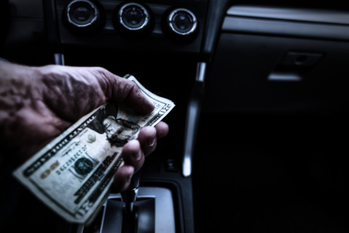 A shady character con man in the driver's seat of a darkened and somewhat grubby car interior is offering a stack of money to an unseen potential passenger. Payoff? Soliciting? Bribing? Money laundering? Illicit temptation? Other criminal or underground economic activity?
