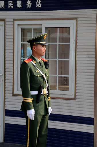 Beijing, China - October 18, 2015: A Chinese police officer stands smartly at attention on the outskirts of Tiananmen Square, Beijing, China. He wears his green uniform with gold braids and white gloves. Behind him is a small police station. 