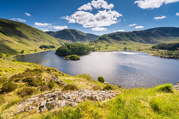 Sunny day at Haweswater reservoir in England Haweswater Reservoir in Mardale Valley taken from Whiteacre Crag. A green hill that leads down to the water is in the foreground. The water is deep blue and reflects the sunlight. The reservoir is surrounded by rolling green hills, and a deep blue sky filled with fluffy white clouds is overhead. reservoir stock pictures, royalty-free photos & images