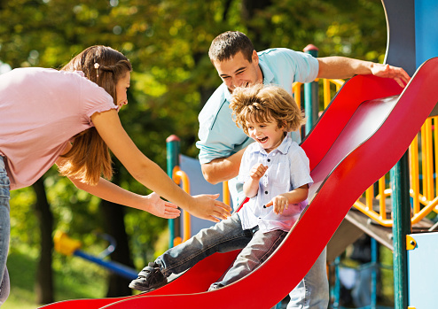 Cheerful parents having fun with their son in the playground. Little boy is sliding.   