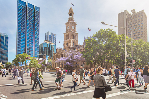 People walking in George St on a sunny day with the Sydney Town Hall building in the background.
