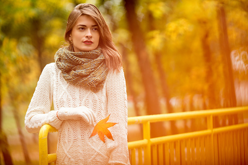 Beautiful young woman enjoy in the park covered with golden automn leaves. She is relaxing on the yellow bridge and she pensive looking into the distance and smiling. She's wearing a handmade wool scarf, sweater and holiding one leaf. 