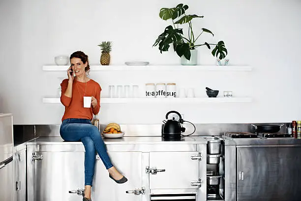Shot of a woman sitting on her kitchen counter drinking a coffee and talking on the phone