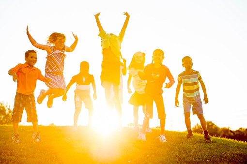 A multi-ethnic group of elementary age children ares jumping on a hill top on a beautiful sunny day.