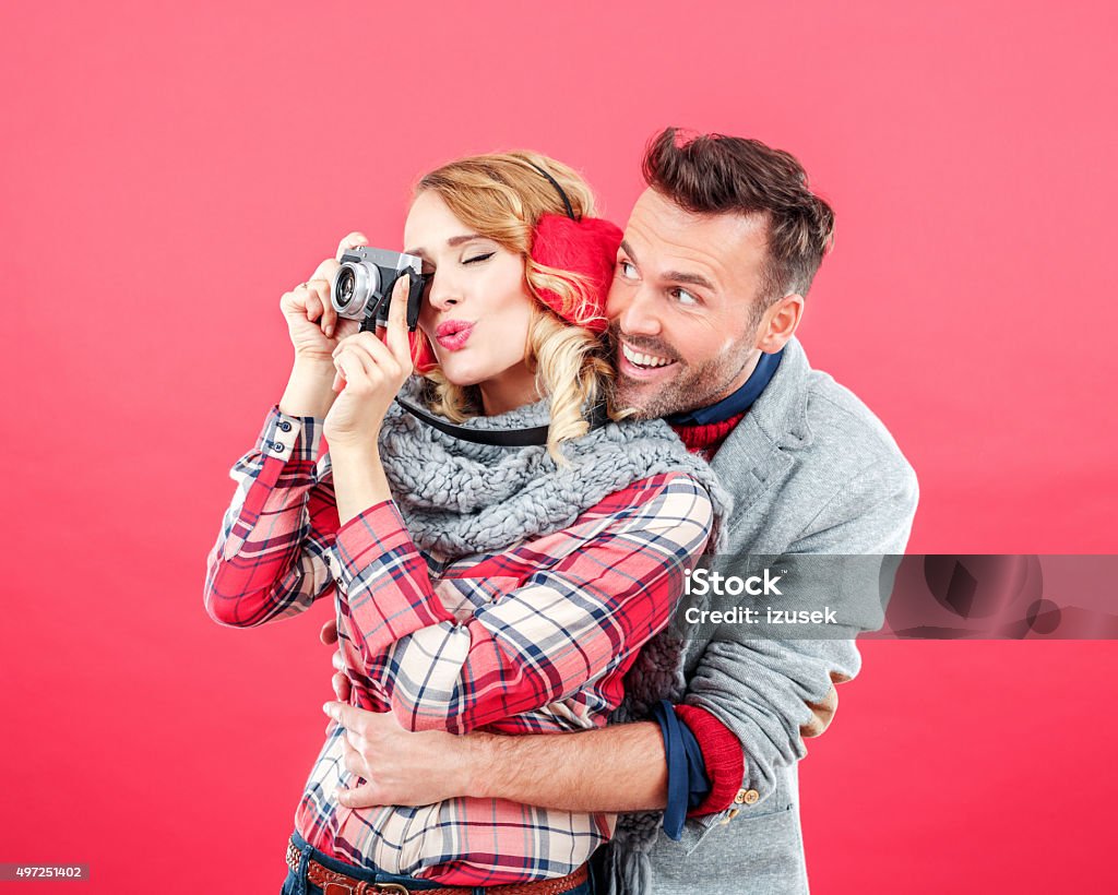 Happy couple in winter outfit against red background Studio portrait of happy, affactionate couple in winter outfits standing against red background, woman holding camera and photographing. 2015 Stock Photo