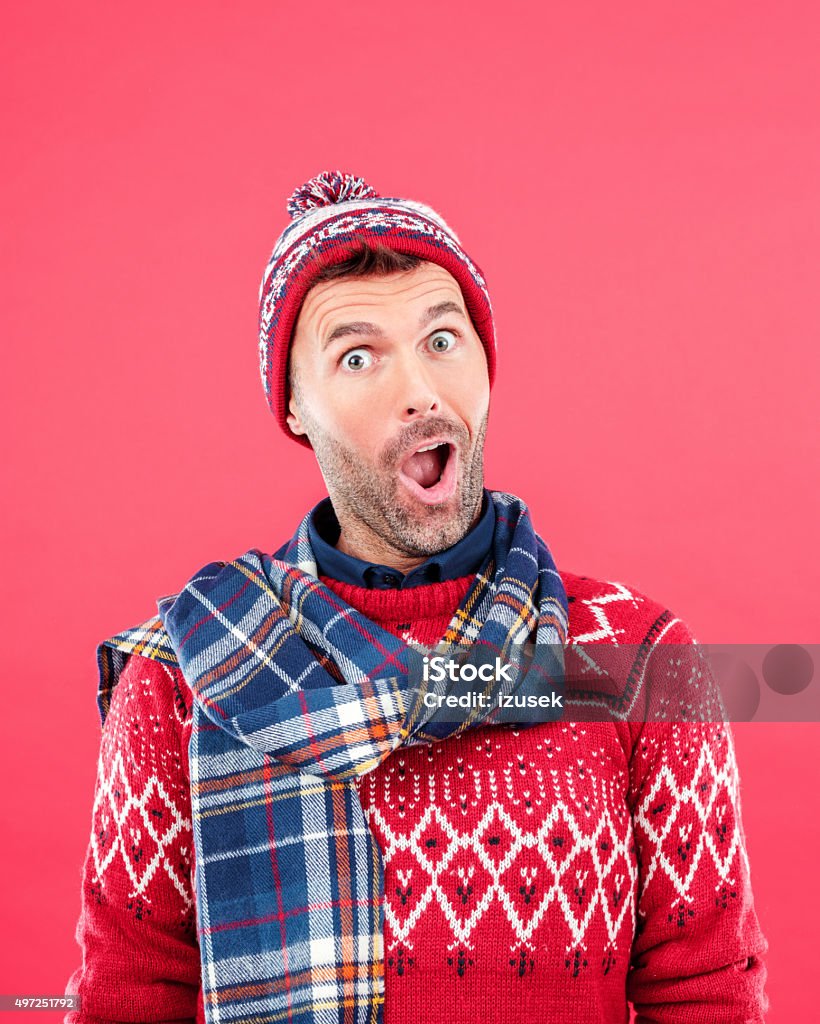 Excited man in winter outfit against red background Studio portrait of happy man in winter outfit - cap, scarf and sweater, standing against red background and staring at camera with mouth open, rolling his eyes. Men Stock Photo