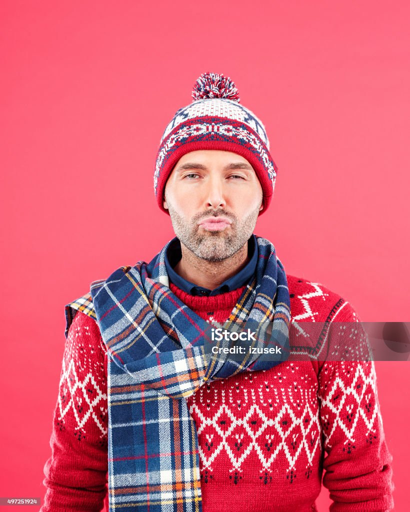 Cute man in winter outfit against red background Studio portrait of happy man in winter outfit - cap, scarf and sweater, standing against red background and kissing. Men Stock Photo