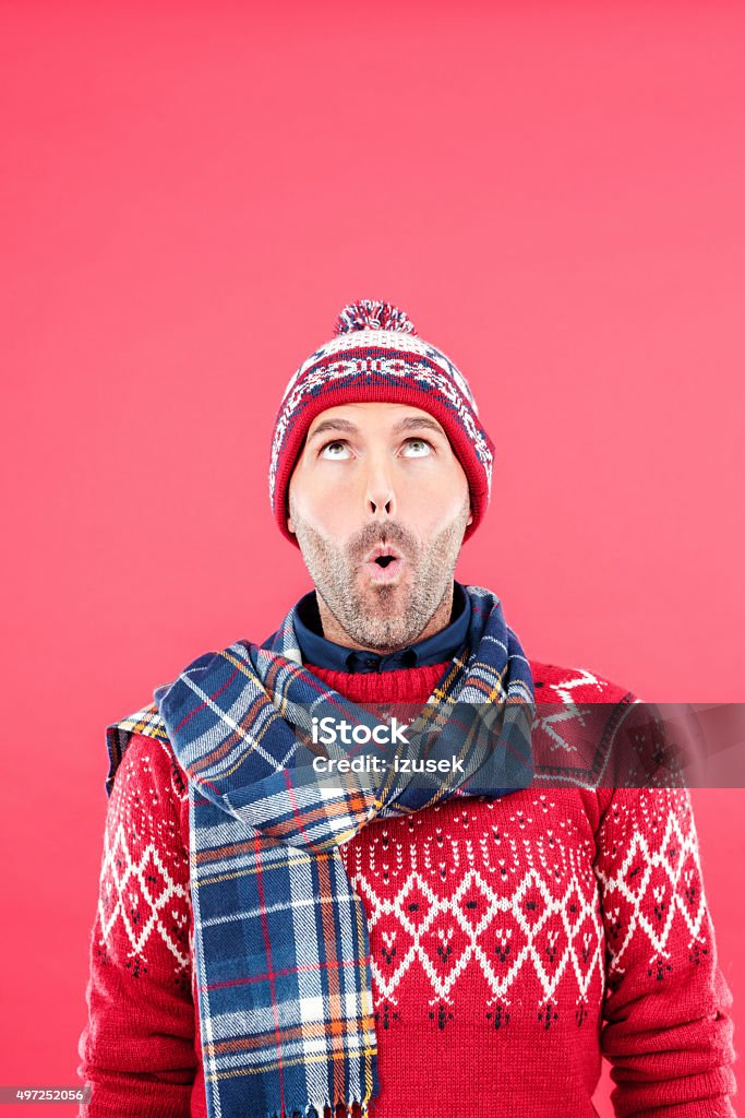 Surprised man in winter outfit against red background Studio portrait of surprised man in winter outfit - cap, scarf and sweater, standing against red background, looking up. Portrait Stock Photo