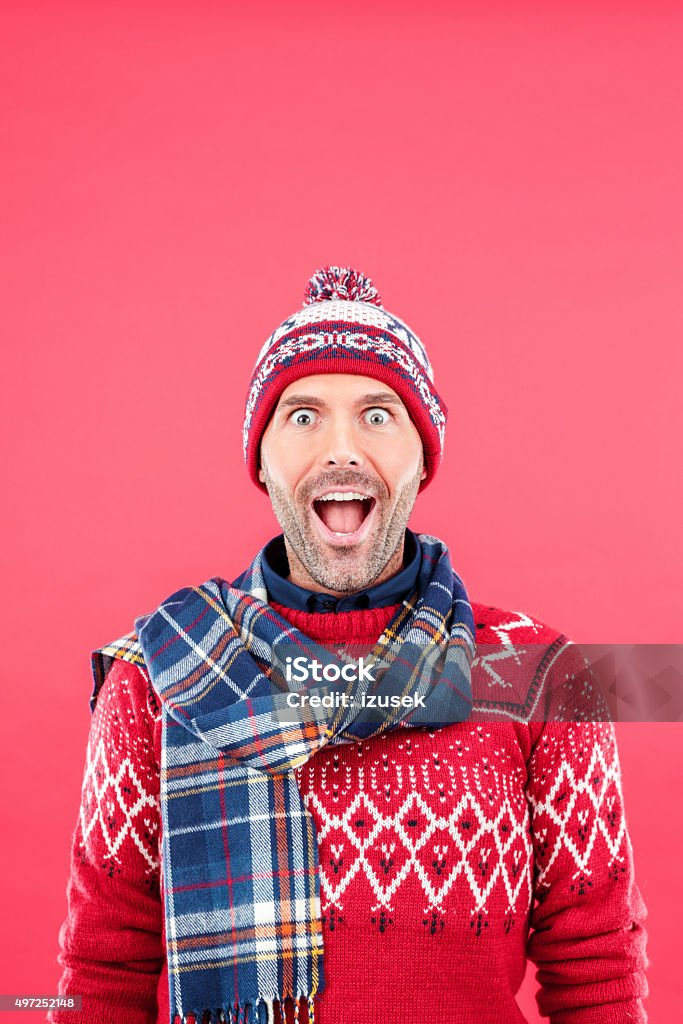 Excited man in winter outfit against red background Studio portrait of surprised man in winter outfit - cap, scarf and sweater, standing against red background, staring at camera with mouth open, rolling his eyes. Christmas Stock Photo
