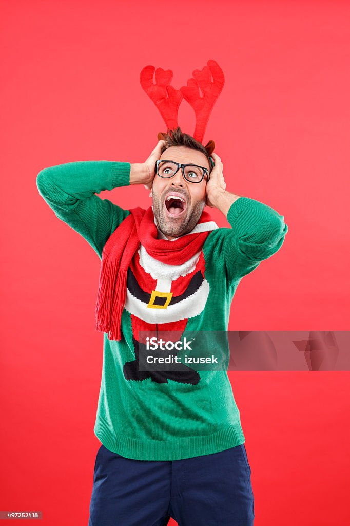 Terrified nerd man in funny winter outfit against red background Studio portrait of terrified man wearing santa christmas sweater and reindeer antlers headband, standing against red background, shouting, rolling eyes, holding head in hands. Christmas Sweater Stock Photo