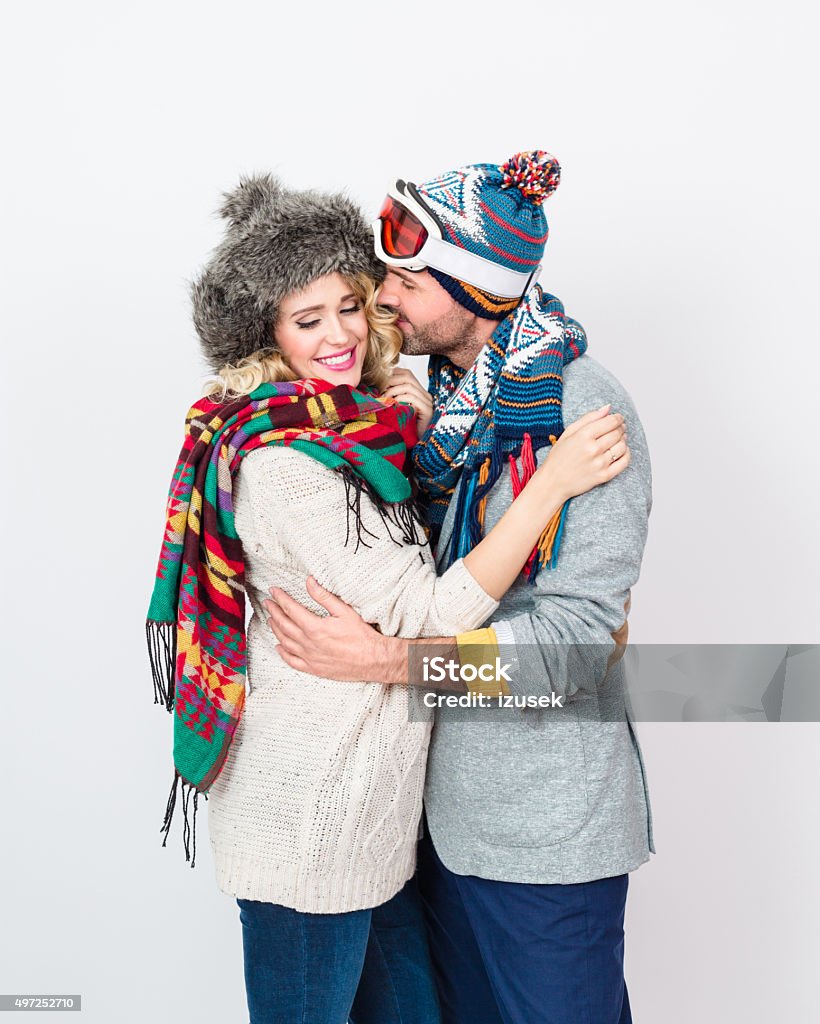 Affectionate couple in winter outfit against white background Studio portrait of happy, affactionate couple in winter outfits - caps, scarfs, sweaters, embracing against white background. 2015 Stock Photo
