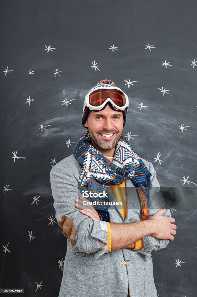 Happy man in winter outfit against blackboard Studio portrait of handsome man in winter outfit - cap, scarf and goggles, standing against blackboard decorated with snowflakes, laughing at camera. Arms Crossed Stock Photo