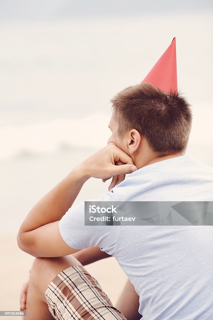 Lonely birthday beach party Retro colored back view portrait of young man wearing red party hat, blues t-shirt and checkered shorts celebrating birthday sitting alone with hand under his chin on beach near tropical sea Beach Stock Photo