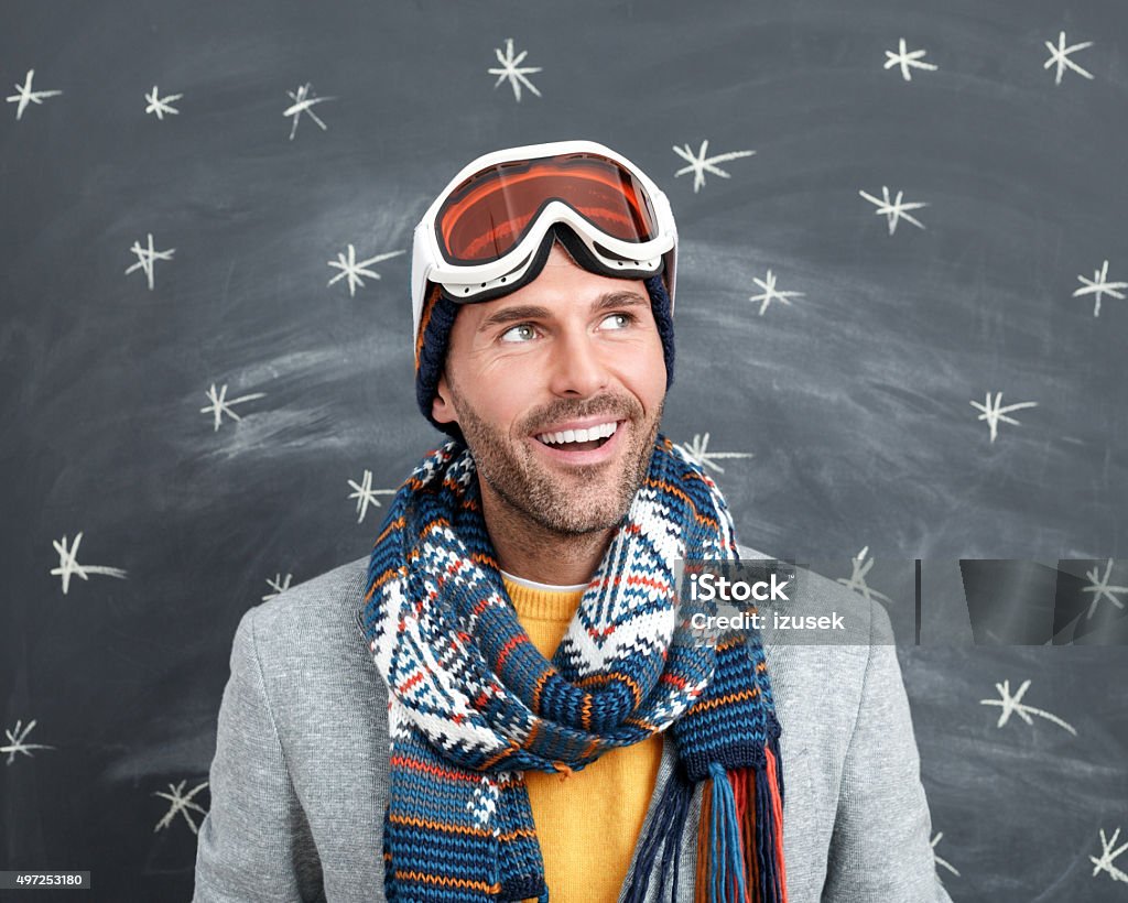 Happy man in winter outfit against blackboard Studio portrait of handsome man in winter outfit - cap, scarf and goggles, standing against blackboard decorated with snowflakes. Close up of face. 2015 Stock Photo