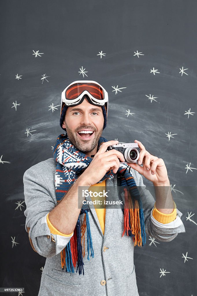 Happy man in winter outfit against blackboard, holding a camera Studio portrait of handsome man in winter outfit - cap, scarf and goggles, standing against blackboard decorated with snowflakes, holding camera in hands and laughing. Camera - Photographic Equipment Stock Photo