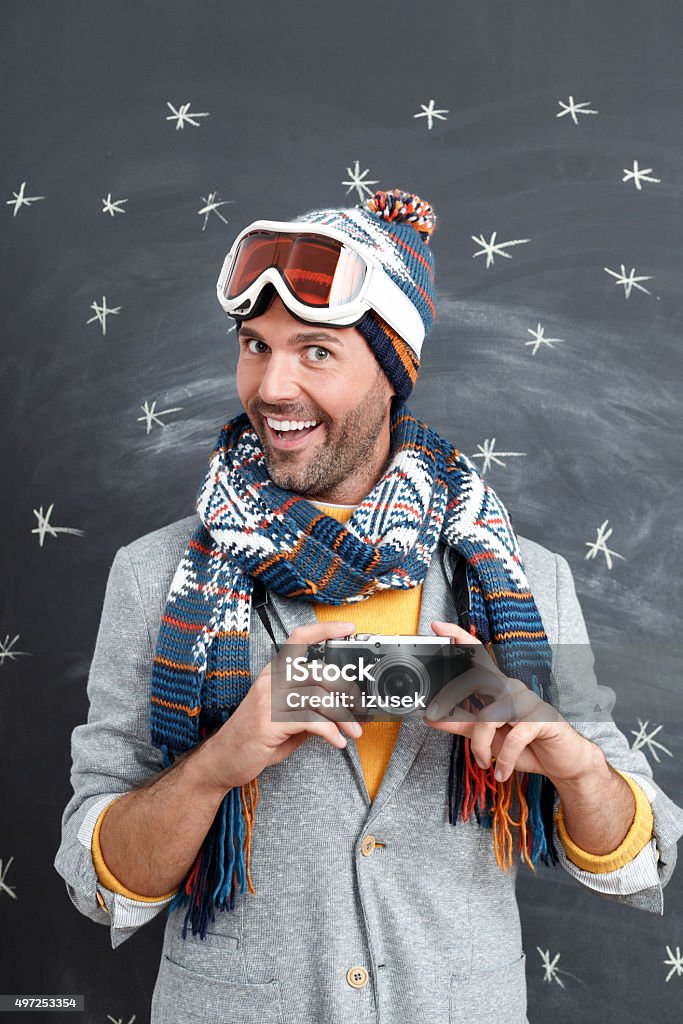Excited man in winter outfit against blackboard, holding a camera Studio portrait of handsome man in winter outfit - cap, scarf and goggles, standing against blackboard decorated with snowflakes, holding camera in hands and laughing. Men Stock Photo