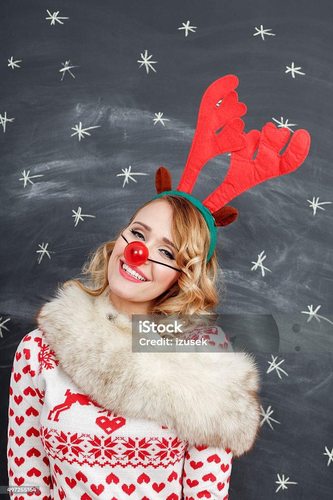 Happy woman in winter outfit and reindeer antlers headband Studio portrait of happy young woman in winter outfit - christmas sweater, fur shawl, gloves, reindeer antlers headband and red nose standing against blackboard decorated with snowflakes. 2015 Stock Photo