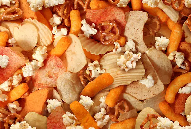 Snacks background with salty crunchy treats as potato chips and cheese flavored puffs fried or baked food as pretzels pop corn and nachos as a symbol of assorted party mix appetizer.